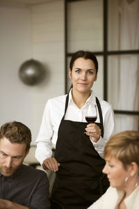 Portrait of woman wearing apron holding wineglass while standing by people