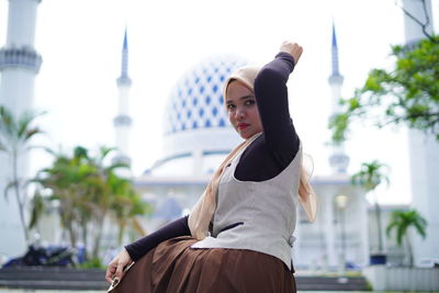 Young woman looking away while sitting on staircase against mosque