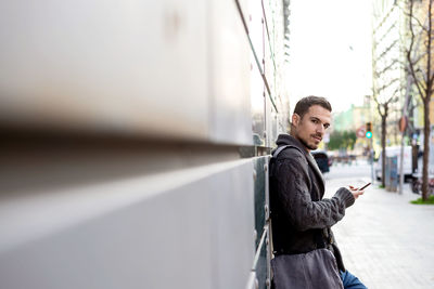 Side view of a bearded man using phone leaning on office building wall