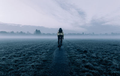 Rear view of man cycling on field against sky during foggy weather