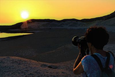Woman photographing at beach against sky during sunset