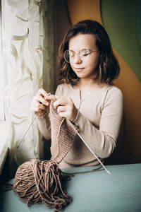Teenage girl in a beige sweater, wearing glasses knitting needles standing by the window 
