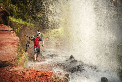 Man carrying mountain bike and walking by waterfall