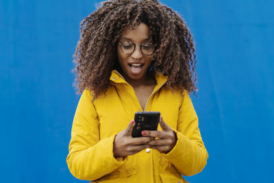 Young woman using phone while standing against blue sky