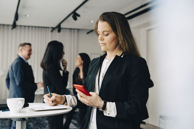 Female business person using phone and writing while standing at table in office workplace