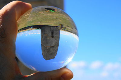 Cropped hand holding crystal ball with reflection on tower against blue sky