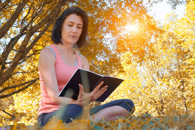 Beautiful young woman using smart phone while sitting on autumn leaves
