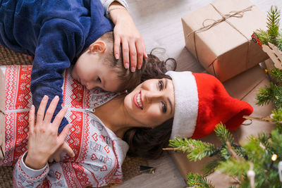 Happy mom with son in santa hat lie on floor at home under tree with gifts