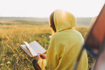 Rear view of woman wearing hood reading book on field