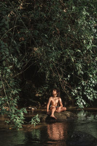 Positive boy in swimming trunks sitting on stone on calm river coast in lush woodland and looking away in dreams