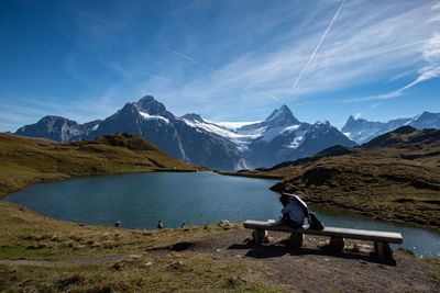 Man sitting on bench by sea against snowcapped mountains