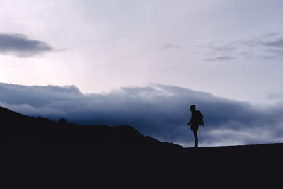 Side view silhouette of woman with backpack standing on glade with camera in magnificent valley against foggy ridges at horizon under sky with fluffy clouds in spain