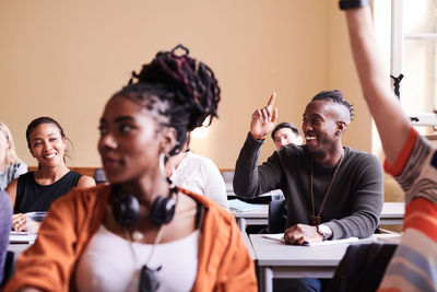 Happy young male student raising hand while sitting with classmates in school building