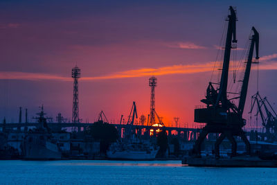 Cranes at harbor against sky during sunset