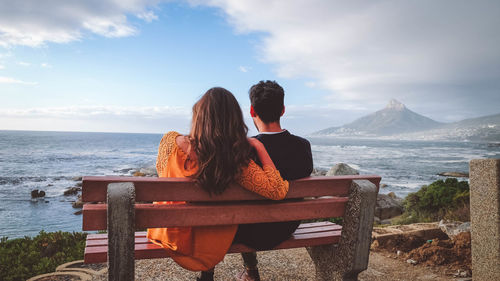 Rear view of couple sitting on bench while looking at sea against sky