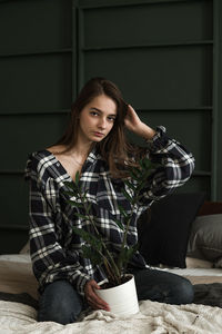Portrait of young woman with potted plant kneeling on bed against wall at home