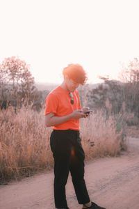 Full length of man using mobile phone on dirt road by plants against sky