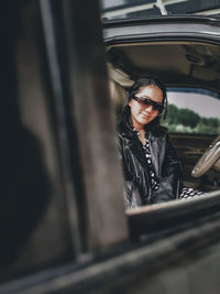 A girl wearing a polkadot dress, jacket, sun glasses and high boots sitting in a car 