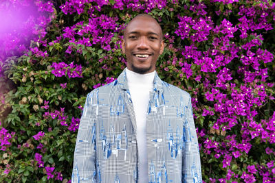 Delighted african american male in stylish outfit looking at camera while standing near bush with blooming flowers on summer day