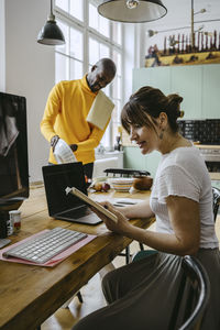 Mid adult woman sitting with laptop reading book while boyfriend doing chores standing at table