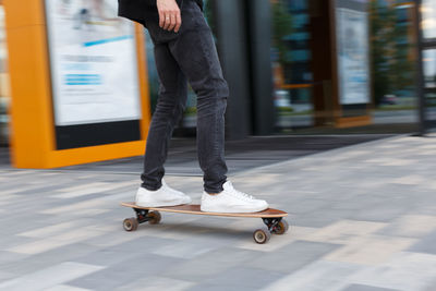 Shot of male in black jeans and white sneakers riding a longboard in urban area, photo in motion