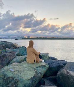 Rear view of woman sitting on rock by sea against sky