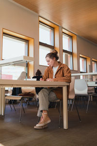 Young woman in casual clothes with hair bun writing in planner while sitting at table and studying in university library
