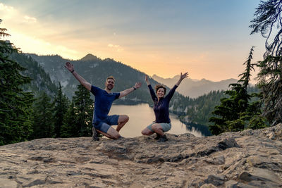 Portrait of couple with arms outstretched crouching on mountain against sky during sunset