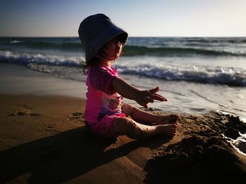 Boy gesturing while sitting at beach against sky 