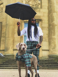 Man with dog holding umbrella while standing on road against building