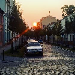 Cars on city street by buildings against sky during sunset