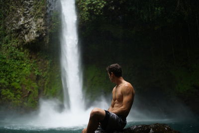 Shirtless man sitting in front of la fortuna waterfall in costa rica