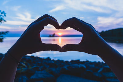 Close-up of hand holding heart shape against sky during sunset