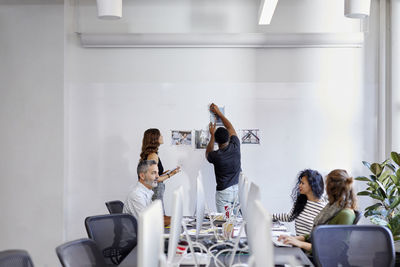 Business people discussing at conference table while colleagues attaching photograph printouts on bulletin board in office