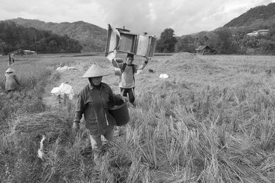 People carrying objects while walking on grassy field against sky