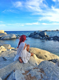 Rear view of woman sitting on rock by sea against sky