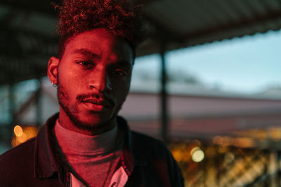 Young african american hipster male with curly hair and beard wearing warm jacket looking at camera while standing against shabby wall