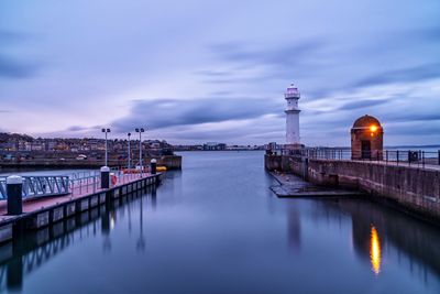 Illuminated bridge over river by buildings against sky at dusk