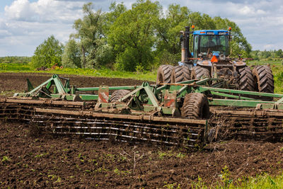Blue tractor with double wheels pulling disc harrow with roller basket at hot sunny day