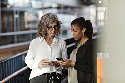 Female business colleagues talking while using smart phones at office lobby