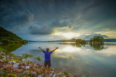 Rear view of man with arms raised standing by lake against sky during sunset