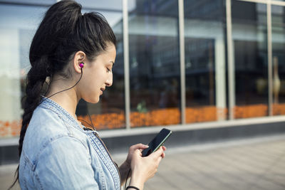 Side view of woman listening music in mobile phone while standing in city