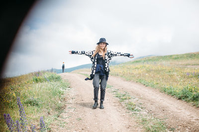 Full length of woman with arms outstretched standing amidst grassy field on dirt road