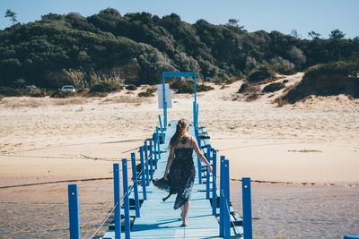 Rear view of young woman walking on pier at beach against sky