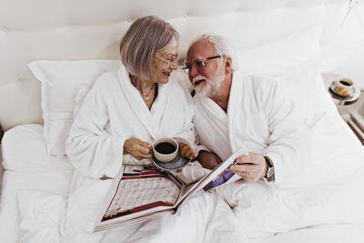 Senior woman looking at man holding newspaper while having coffee on bed in hotel