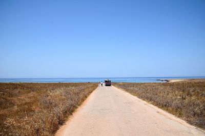 Road amidst field leading towards sea against clear blue sky