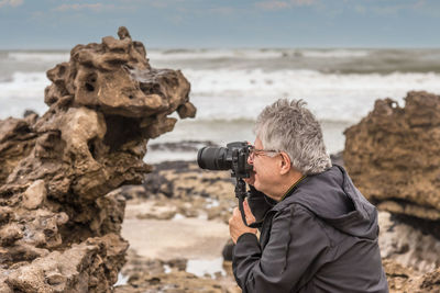 Gray haired mature adult man with glasses taking a photograph near the sea coast.