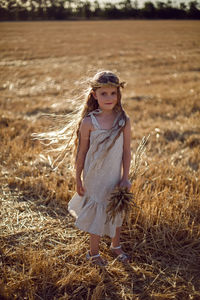 Girl child in a dress and a wreath on her head stands on a mown field of wheat at sunset in summer