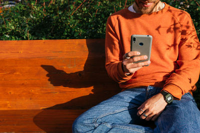 Cropped unrecognizable male in cap surfing internet on cellphone while sitting on bench in park near green plants on sunny street