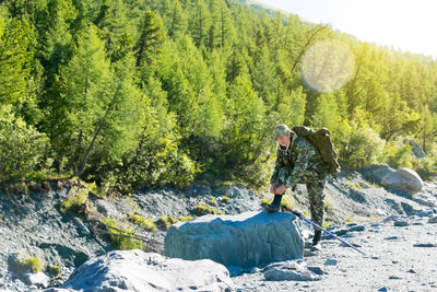 Portrait of backpack man tying shoelace on rock by trees at forest